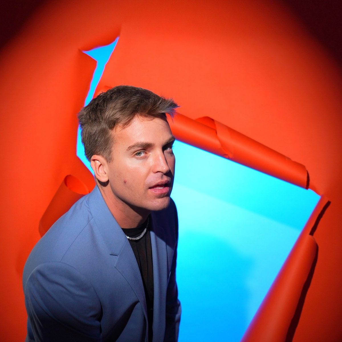 James Maas, host of The Daily Skinny, in a blue blazer, leaning out of a torn orange wall with bright blue behind it